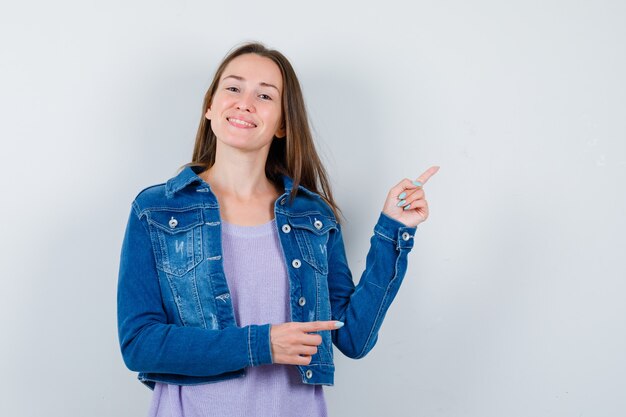 Giovane donna che indica nell'angolo in alto a destra in t-shirt, giacca e sembra allegra. vista frontale.