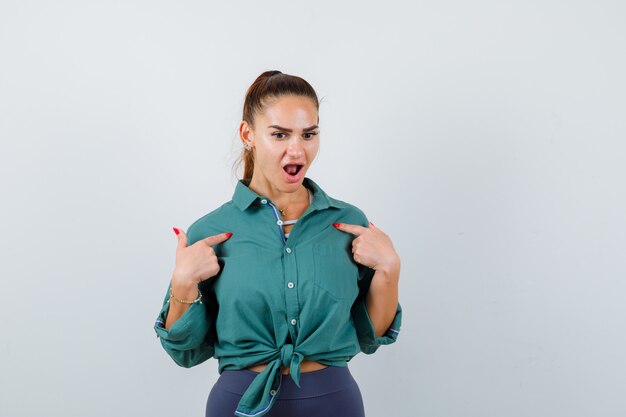 Giovane donna che indica se stessa in camicia verde e guarda domandata, vista frontale.