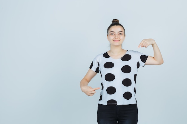 Giovane donna che indica se stessa in t-shirt, jeans e guardando orgoglioso, vista frontale.