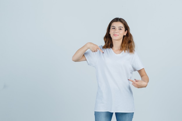 Giovane donna che indica se stessa in t-shirt, jeans e guardando orgoglioso, vista frontale.