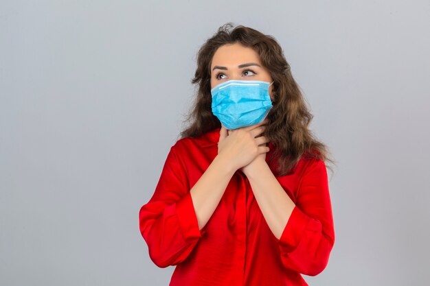 Giovane donna che indossa camicetta rossa in mascherina protettiva medica che sembra malata toccando il suo collo e soffre di mal di gola su sfondo bianco isolato