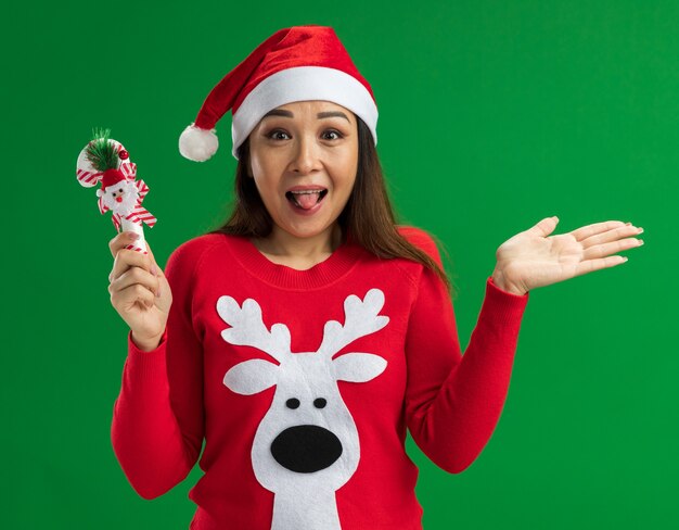 Giovane donna che indossa il cappello della santa di Natale e maglione rosso che tiene il bastoncino di zucchero di Natale che guarda l'obbiettivo felice ed eccitato che attacca fuori la lingua che si leva in piedi sopra la priorità bassa verde