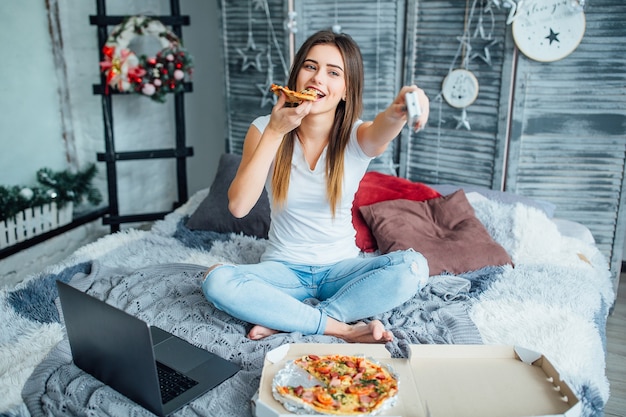 Giovane donna che indossa in stile casual seduta sul letto con pizza e laptop e accende la TV.