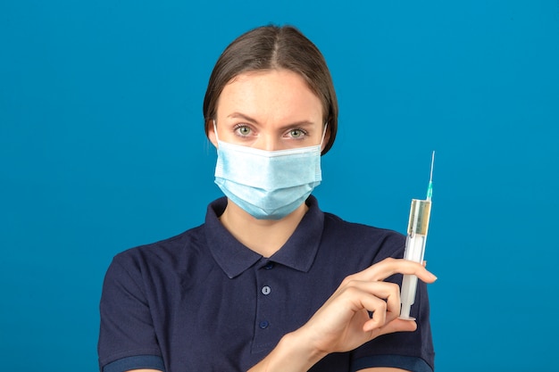 Giovane donna che indossa la camicia di polo blu in siringa protettiva della tenuta della maschera medica che esamina macchina fotografica con il fronte serio che sta sul fondo blu isolato