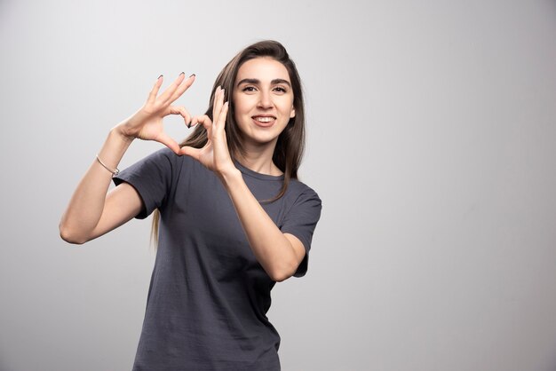 Giovane donna che indossa la maglietta grigia su sfondo grigio facendo forma di simbolo del cuore con le mani.