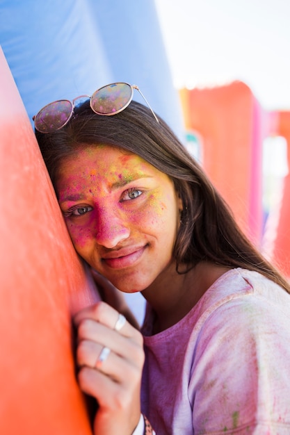 Giovane donna che indossa occhiali da sole con il viso coperto di colore holi rivolto verso l&#39;obiettivo