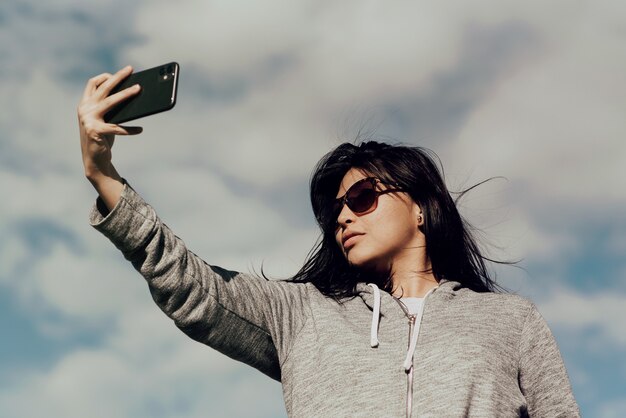 Giovane donna che indossa occhiali da sole di scattare una foto con il suo telefono sotto il cielo blu nuvoloso