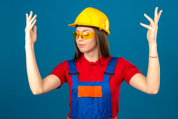 Giovane donna che indossa occhiali gialli in uniforme di costruzione e casco di sicurezza giallo in piedi con gli occhi chiusi e le mani sollevate su sfondo blu