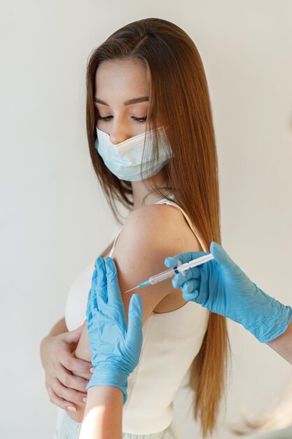Giovane donna che indossa una maschera e riceve il vaccino contro il coronavirus Una giovane ragazza che riceve un'immunità colpita all'avambraccio Primo piano del medico con guanti blu che danno Covid19 AIDS