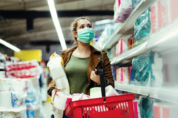 Giovane donna che indossa una maschera facciale e fa forniture di carta igienica mentre acquista al supermercato in tempo di pandemia di virus