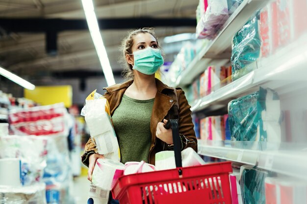 Giovane donna che indossa una maschera facciale e fa forniture di carta igienica mentre acquista al supermercato in tempo di pandemia di virus
