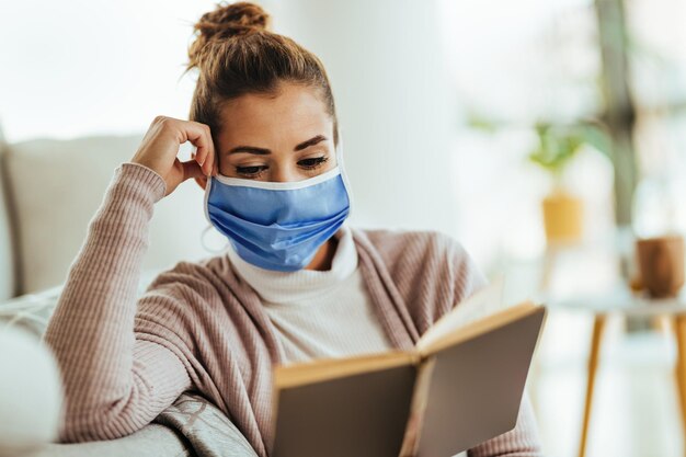 Giovane donna che indossa una maschera per il viso mentre si rilassa a casa e legge un libro