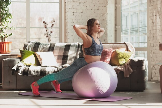 Giovane donna che insegna a casa corsi online di fitness, aerobica, stile di vita sportivo durante la quarantena. Diventare attivi mentre si è isolati, benessere, concetto di movimento. Esercizi con fitball per la parte inferiore del corpo.