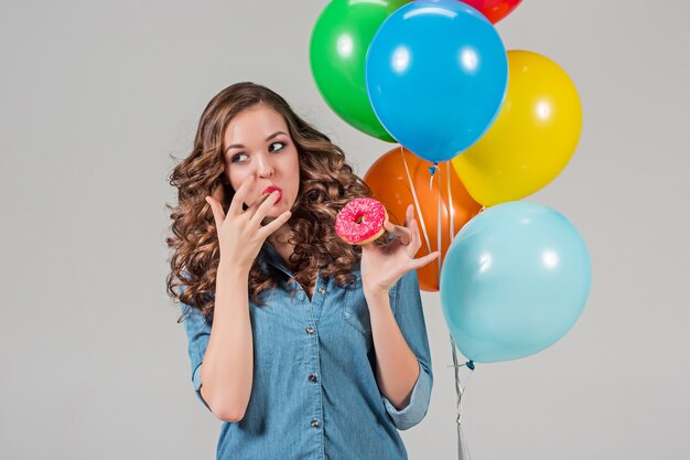 Giovane donna che mangia ciambella e che tiene palloncini colorati sulla parete grigia dello studio