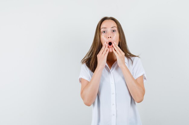 Giovane donna che mette le mani sul mento in camicetta bianca e sembra sbalordita.