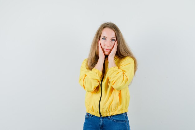 Giovane donna che mette le mani sulla guancia in bomber giallo e jeans blu e guardando affascinante, vista frontale.