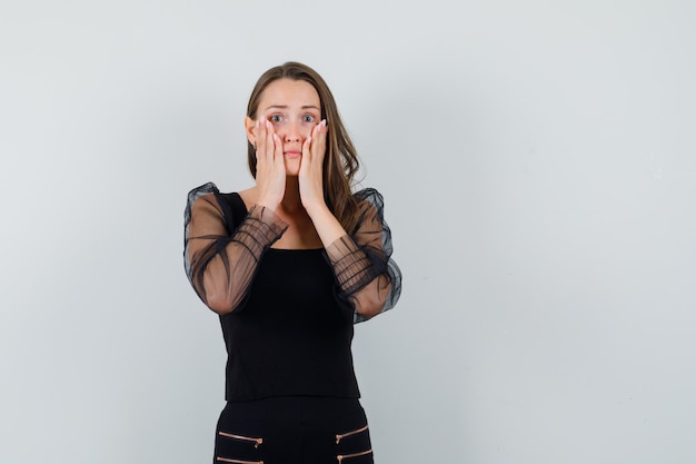 Giovane donna che mette le mani sulle guance in camicetta nera e pantaloni neri e sembra timida, vista frontale.