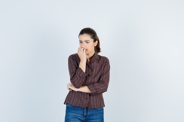 Giovane donna che morde il pugno emotivamente in camicia a righe, jeans e sembra ansiosa, vista frontale.