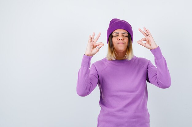 Giovane donna che mostra gesto di meditazione in maglione viola, berretto e dall'aspetto pacifico, vista frontale.