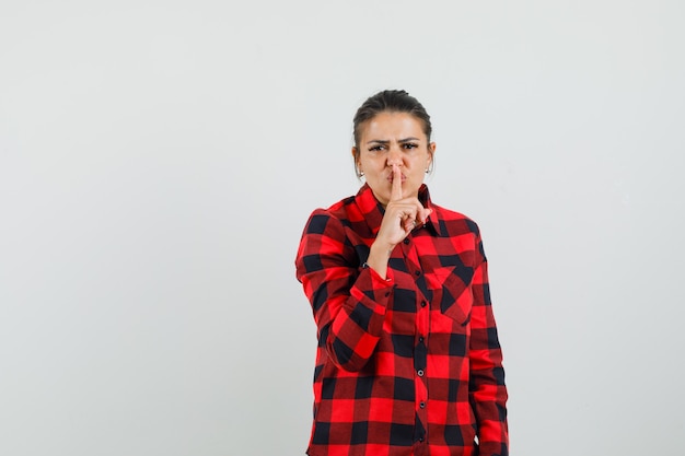 Giovane donna che mostra gesto di silenzio in camicia a quadri e guardando attento, vista frontale.