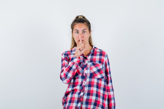 Giovane donna che mostra gesto di silenzio in camicia casual e guardando attento. vista frontale.