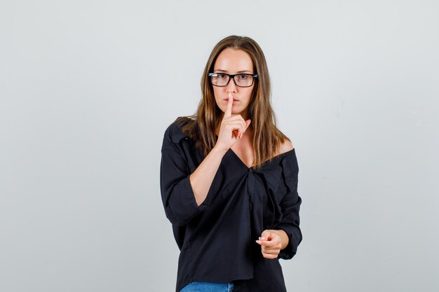 Giovane donna che mostra gesto di silenzio in camicia, pantaloncini, occhiali e guardando attento. vista frontale.