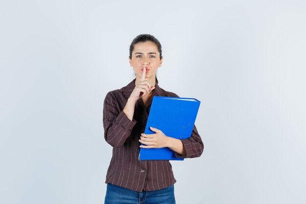 Giovane donna che mostra gesto di silenzio, tenendo la cartella in camicia, jeans e guardando sensata, vista frontale.