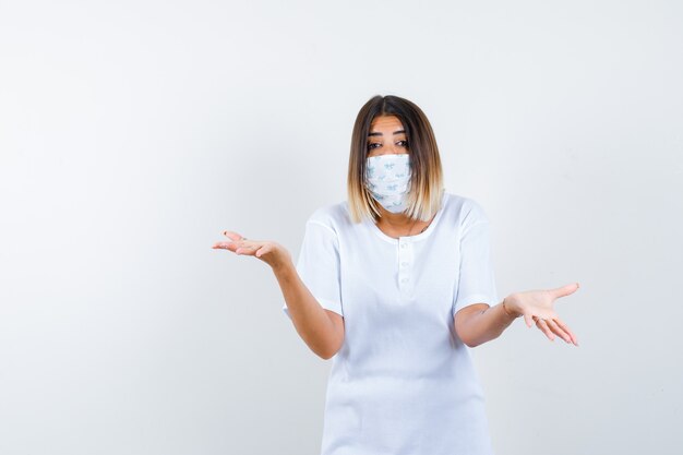 Giovane donna che mostra gesto impotente in maglietta, maschera e sguardo stressato, vista frontale.