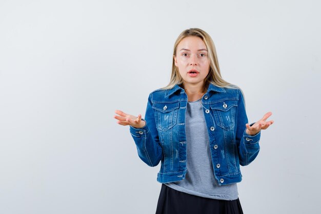 Giovane donna che mostra gesto impotente in t-shirt, giacca di jeans, gonna e sembra stupita.