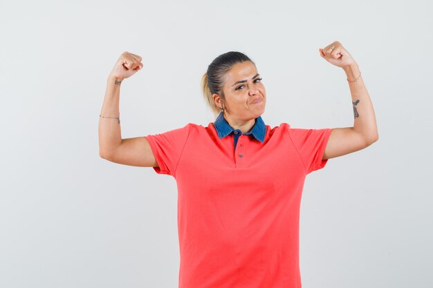 Giovane donna che mostra i muscoli in maglietta rossa e guardando fiducioso, vista frontale.