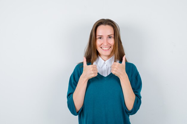 Giovane donna che mostra i pollici doppi in maglione sopra la camicia bianca e sembra felice, vista frontale.