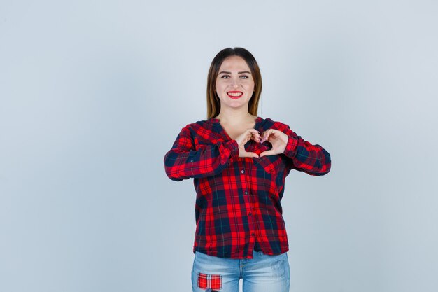 Giovane donna che mostra il gesto del cuore in camicia a quadri e sembra allegra. vista frontale.