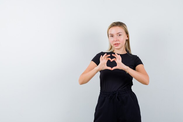 Giovane donna che mostra il gesto del cuore in t-shirt, pantaloni e carino, vista frontale.