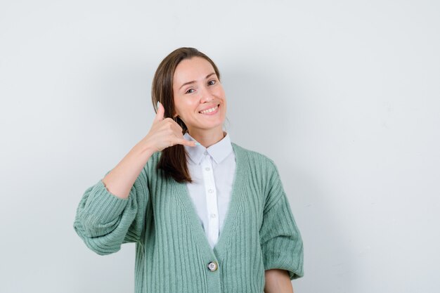 Giovane donna che mostra il gesto del telefono in camicetta, cardigan e sembra allegra. vista frontale.