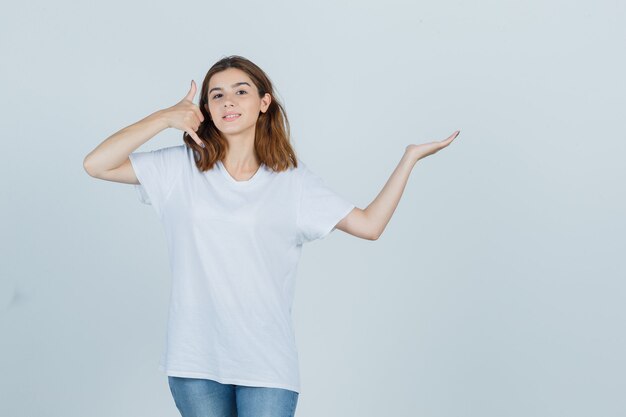 Giovane donna che mostra il gesto del telefono mentre finge di tenere qualcosa in t-shirt, jeans e sembra sicura, vista frontale.