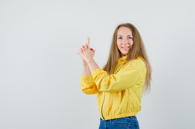 Giovane donna che mostra il gesto della pistola in bomber giallo e jeans blu e guardando ottimista, vista frontale.