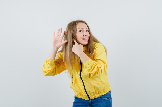 Giovane donna che mostra il gesto di ascolto in bomber giallo e jeans blu e sembra ottimista. vista frontale.