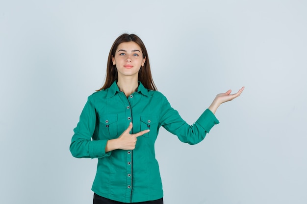 Giovane donna che mostra il gesto di benvenuto mentre indica da parte in camicia verde e sembra fiduciosa, vista frontale.