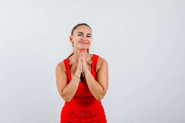 Giovane donna che mostra il gesto di namaste in canottiera rossa, pantaloni e cerca pacifica, vista frontale.