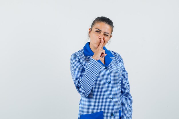 Giovane donna che mostra il gesto di silenzio e mette il dito indice sul labbro in camicia del pigiama a quadretti blu e sembra carina, vista frontale.