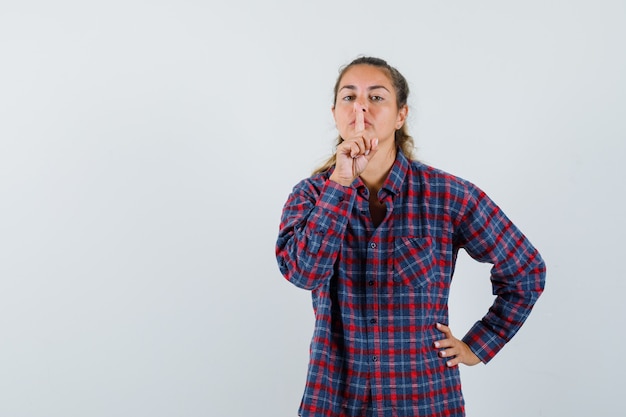 Giovane donna che mostra il gesto di silenzio tenendo la mano sulla vita in camicia a quadri e guardando seria