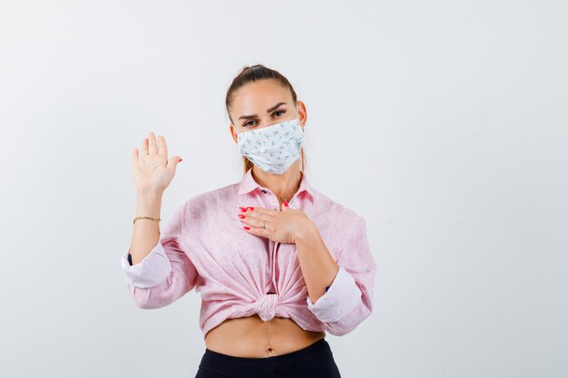 Giovane donna che mostra il palmo, tenendo la mano sul petto in camicia, maschera e guardando allegro, vista frontale.