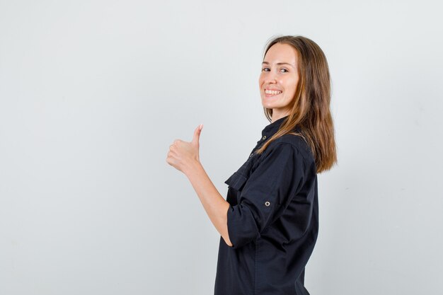 Giovane donna che mostra il pollice in su in camicia nera e sembra allegra. .