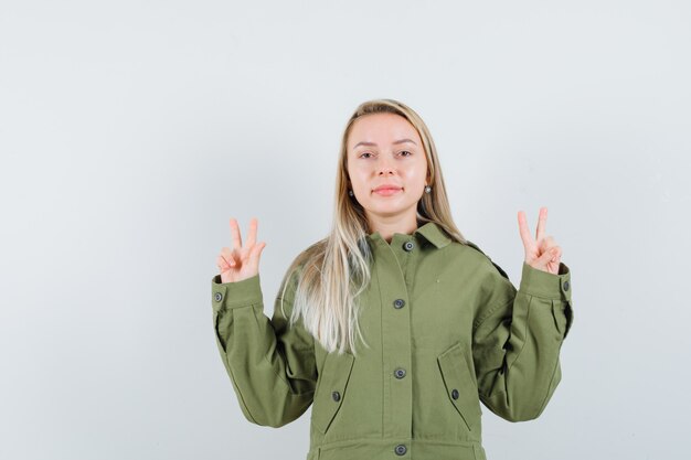 Giovane donna che mostra il segno a V con entrambe le mani in giacca verde, jeans e sguardo ottimista, vista frontale.