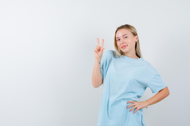 Giovane donna che mostra il segno della vittoria mentre tiene la mano sull'anca in maglietta isolata