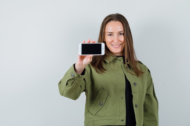 Giovane donna che mostra il telefono cellulare in giacca verde e guardando soddisfatto, vista frontale.