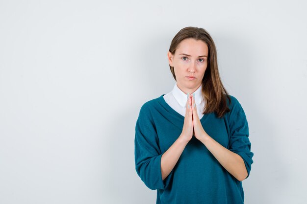 Giovane donna che mostra le mani giunte nel gesto di supplica in maglione sopra la camicia bianca e sembra triste. vista frontale.