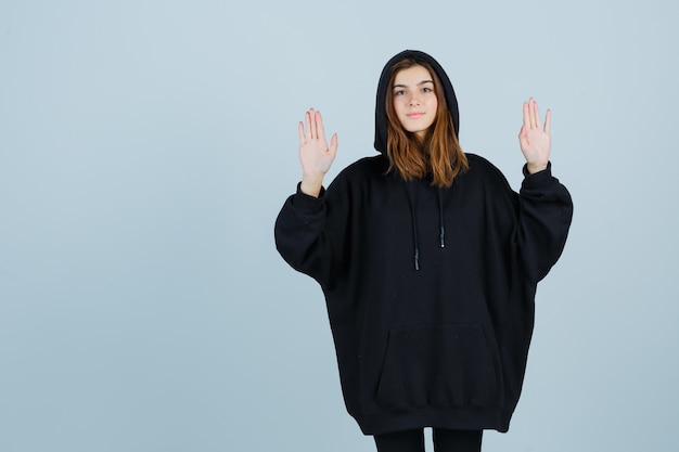 Giovane donna che mostra le palme in gesto di resa in felpa con cappuccio oversize, pantaloni e sembra impotente. vista frontale.