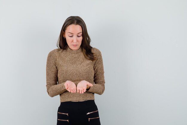 Giovane donna che mostra qualcosa alla sua mano in camicetta dorata e guardando concentrato, vista frontale. spazio per il testo