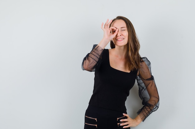 Giovane donna che mostra segno giusto sopra l'occhio e tenendo la mano sulla vita in camicetta nera e pantaloni neri e guardando felice, vista frontale.